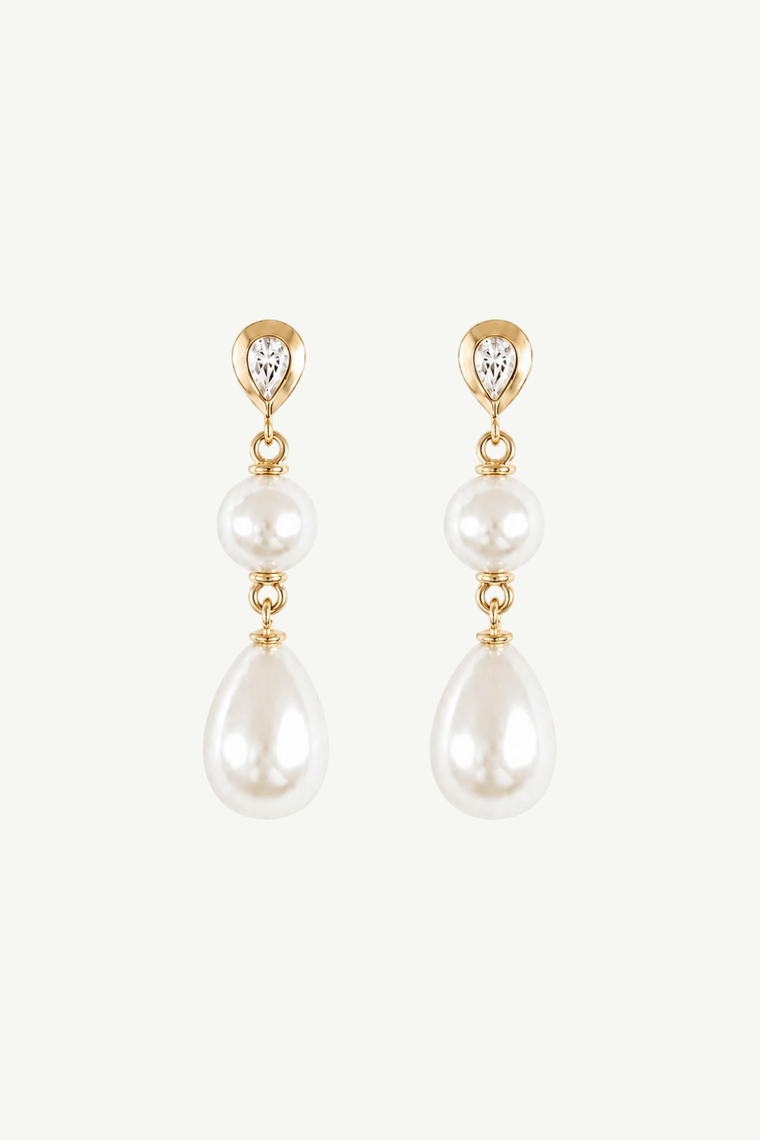 A pair of gold drop earrings featuring a small heart-shaped clear stone at the top, a round pearl, a small gold bead, and a larger teardrop pearl, shown on a light off-white background.